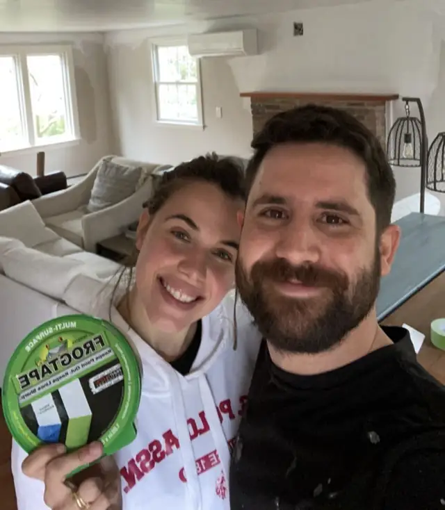 Vini e a esposa numa sala sendo preparada, ela segurando uma fita crepe